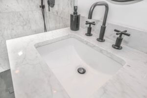 A white sink set in a white marble countertop with stainless steel fixutres.